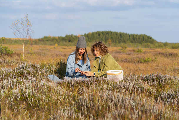 Couple of young women best friends sits sorting berries on autumn meadow grass against blurry green forest. Women wearing blue and green jackets enjoy picnic spending time on nature at sunlight - Foto, Bild