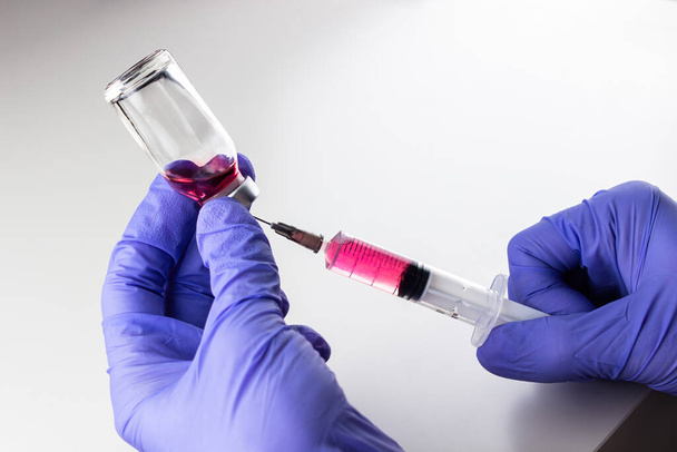 a disposable syringe with a needle and an ampoule with red (vitamin B12) liquid in male hands in blue (lilac) nitrile gloves.  On a white background, lots of empty space - Foto, Bild
