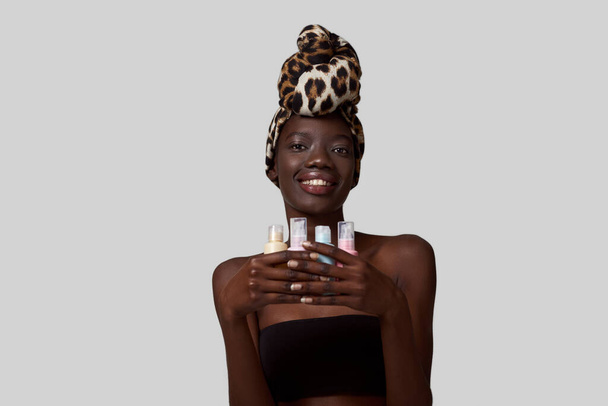 Smiling elegant black girl holding four colorful nail polishes. Beautiful young woman wearing traditional african turban and tank top. Female beauty. White background. Studio shoot. Copy space - Foto, Bild