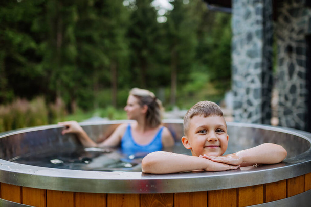 Mother with her little son enjoying bathing in a wooden barrel hot tub in the terrace of the cottage. Wooden bathtub with a fireplace to burn wood and heat water. - Zdjęcie, obraz