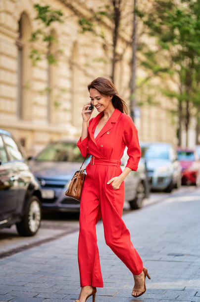 Smiling middle aged woman talking on smartphone. Beautiful female is wearing red clothes on sidewalk at the city. - Foto, Bild