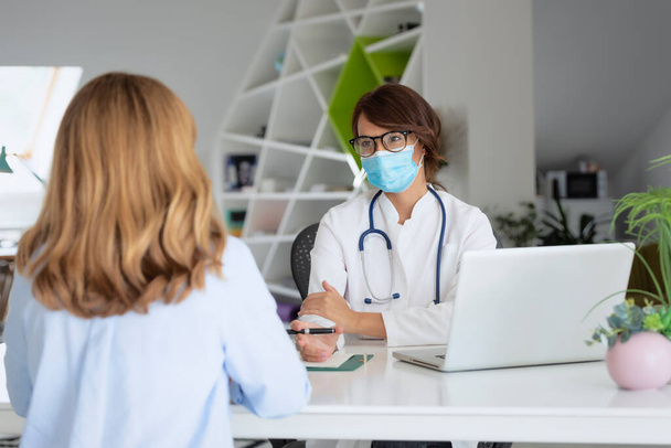 Female doctor wearing face mask while working in the office and listening to the patient. - Foto, afbeelding