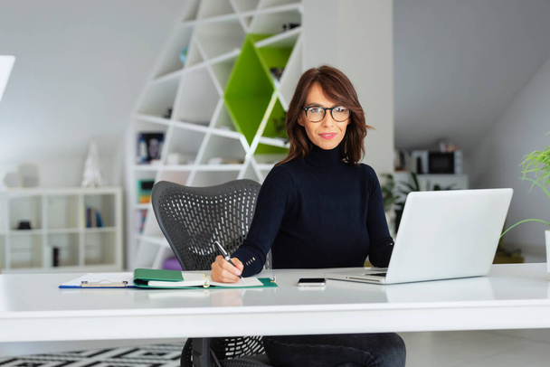 Confident middle aged businesswoman wearing turtleneck sweater while sitting at office desk behind her laptop and working on new project.  - Foto, imagen