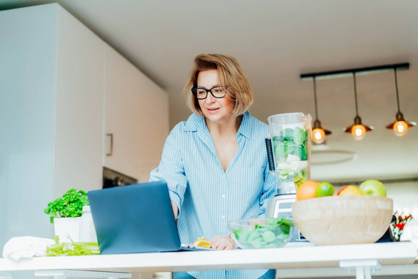 Woman following a cooking tutorial video course on laptop while making green smoothie in a kitchen. Online recipe. Blogger communicates with followers on social media while cooking. Selective focus. - Foto, Bild