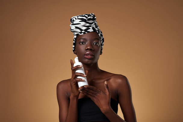 Fashionable black girl holding bottle with cosmetic cream. Pretty young woman wearing traditional african turban and tank top. Body and skin care. Isolated on orange background in studio. Copy space - Foto, immagini