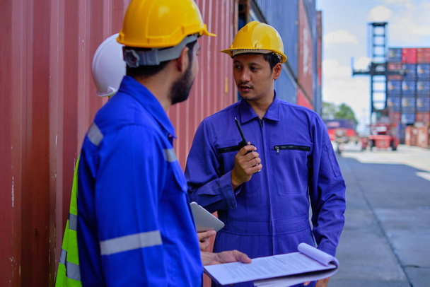 Group of three Asian workers, Thai people in safety uniforms and hard hats work at logistics terminal with many stacks of containers, loading control shipping goods for cargo transportation industry. - Zdjęcie, obraz