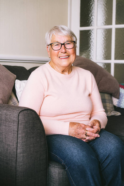 Portrait of smiling senior, mature retired grandmother in eyeglasses, elderly woman smiling sitting on sofa at home enjoying retirement. Vertical card. Selective focus - Valokuva, kuva