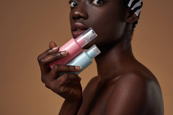 Partial side view of elegant black girl holding colorful nail polishes and looking at camera. Obscure face of youngwoman wear traditional african turban. Orange background. Studio shoot. Copy space - Φωτογραφία, εικόνα