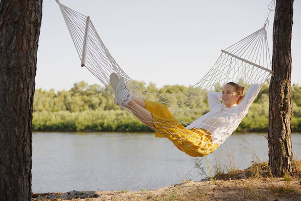 Horizontal shot of positive happy optimistic woman wearing white shirt sitting in hammock on the bank of the river, lying with relaxed expression, looking away with charming smile. - Foto, immagini