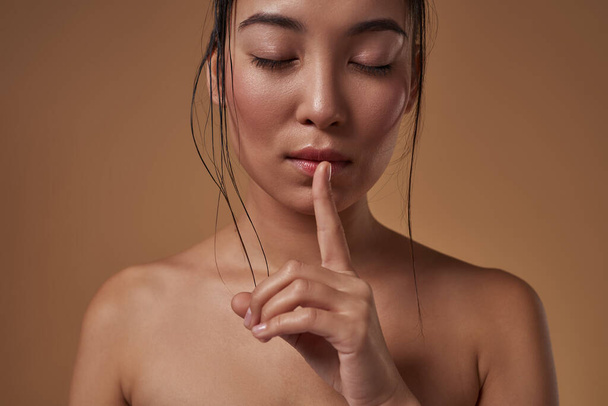 Cropped image of stylish asian girl with closed eyes showing silence sign. Attractive young brunette slim woman. Female beauty. Isolated on orange background. Studio shoot. Copy space - Foto, Imagem