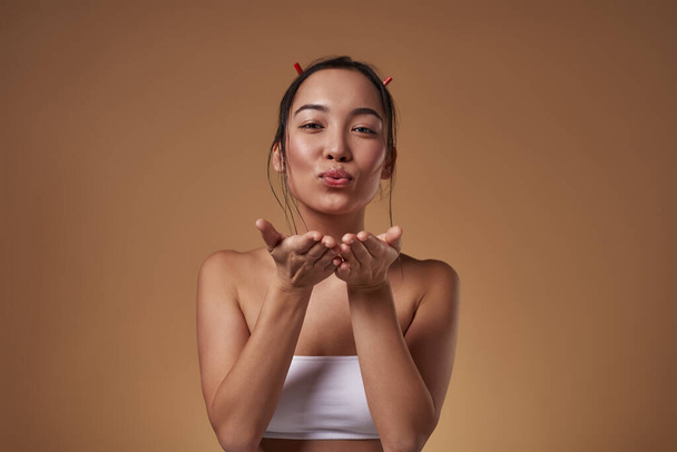 Stylish asian girl blowing air kiss and looking at camera. Attractive young brunette slim woman wearing tank top. Female beauty. Isolated on orange background. Studio shoot. Copy space - Φωτογραφία, εικόνα