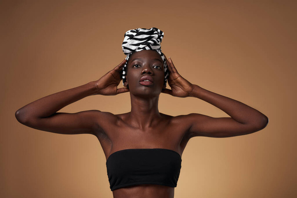 Serious elegant black girl wearing traditional african turban posing and looking at camera. Beautiful young woman wear tank top. Female beauty. Isolated on orange background. Studio shoot. Copy space - Photo, Image