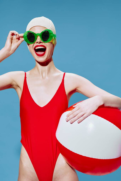 Μια χαρούμενη γυναίκα με κομψά γυαλιά ποζάρει με μια μπάλα κολύμβησης να κοιτάζει την κάμερα. Πορτραίτο μόδας σε μπλε φόντο. Υψηλής ποιότητας φωτογραφία - Φωτογραφία, εικόνα