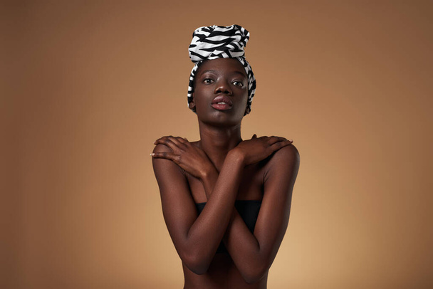 Front view of serious black girl wearing traditional african turban hugging herself. Pretty young slim woman wearing tank top. Female beauty. Isolated on orange background. Studio shoot. Copy space - Photo, Image