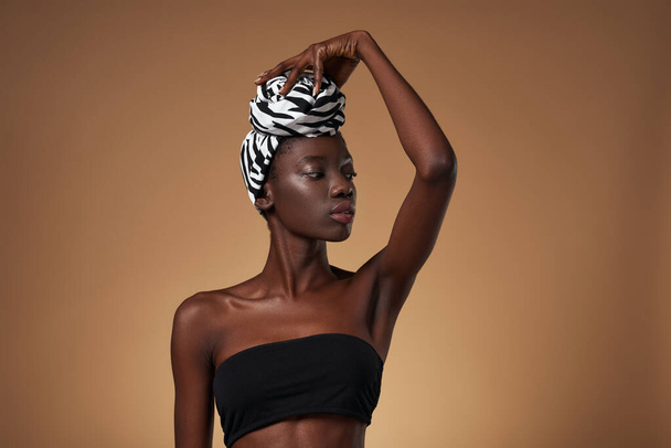 Serious fashionable black girl turban touching traditional african turban on her head. Pretty young slim woman wearing tank top. Orange background. Studio shoot. Copy space - Foto, Imagem