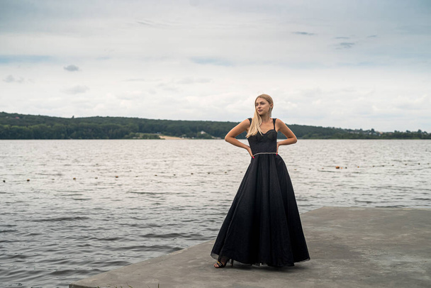 beautiful blonde girl in a black elegant fashionable dress stand near lake, relax and lifestyle - Zdjęcie, obraz