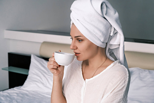 Side view portrait of a young millennial woman drinking morning coffee or tea while sitting on the bed in the bedroom with a white towel on her head in a bathrobe after a shower. Morning concept - Фото, изображение