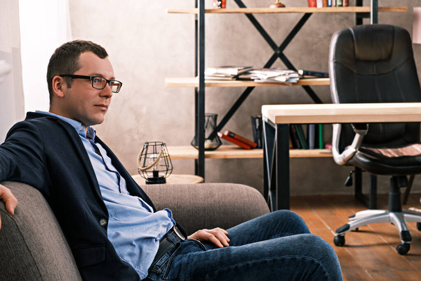 Portrait side view of a Caucasian man in jacket and glasses sitting on sofa looking away against the backdrop of a window and shelves with documents in the office. Success and business concept. - Φωτογραφία, εικόνα