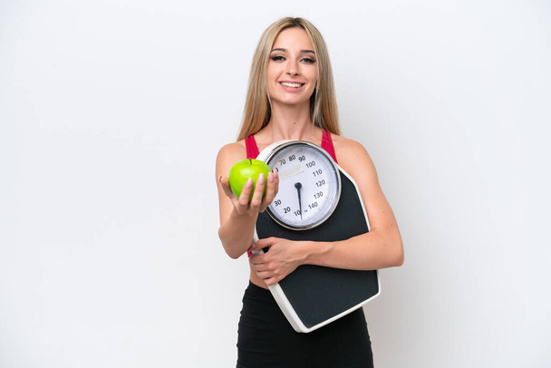 Pretty blonde woman isolated on white background holding weighing machine and offering an apple - Foto, afbeelding