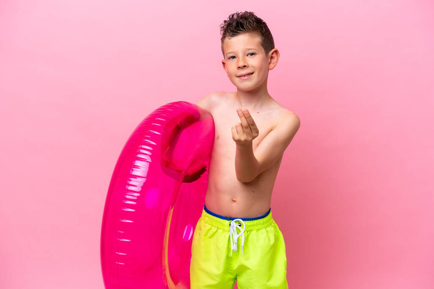 Pequeño chico caucásico sosteniendo un donut inflable aislado sobre fondo rosa haciendo gesto de dinero - Foto, imagen