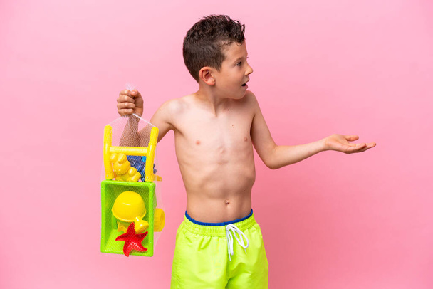 Kleiner kaukasischer Junge hält Strandspielzeug isoliert auf rosa Hintergrund mit überraschendem Gesichtsausdruck - Foto, Bild