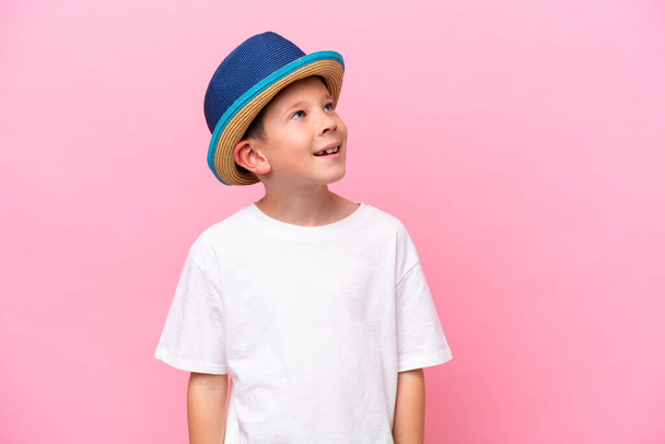 Little caucasian boy wearing a hat isolated on pink background looking up while smiling - Photo, image