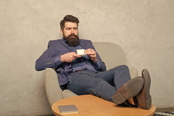 Bearded guy in suit drink tea or coffee relaxing feet up in rest armchair during work break, relax. - Фото, изображение