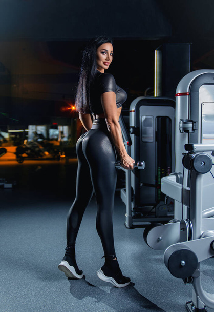 Beautiful fitness brunette girl with perfect back muscles and shapes posing in the gym in sexy black sports costume with dumbbell. - Foto, imagen