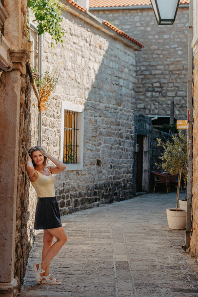 Girl tourist walking through ancient narrow street on a beautiful summer day in MEDITERRANEAN MEDIEVAL CITY , OLD TOWN bUDVA, MONTENEGRO. Young beautiful cheerful woman walking on old street at - Zdjęcie, obraz