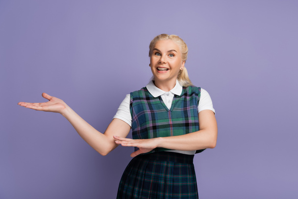 Cheerful student in uniform pointing with hand isolated on purple  - 写真・画像
