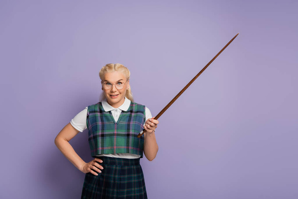 Smiling blonde woman in vest holding pointer and hand on hip on purple background  - Photo, image