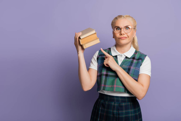 Confused student in eyeglasses pointing at books on purple background  - Fotó, kép
