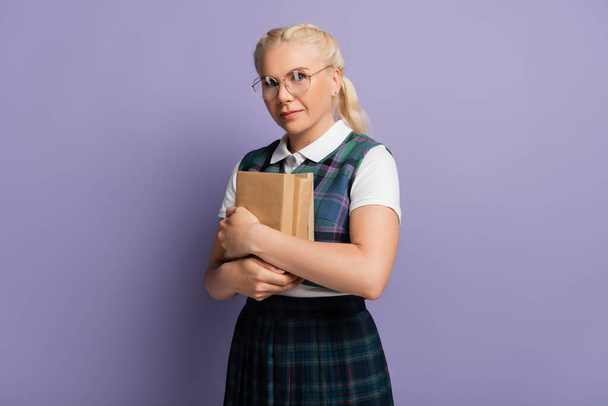 Blonde student in eyeglasses holding books isolated on purple  - 写真・画像