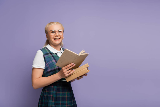 Positive student in eyeglasses and vest holding book isolated on purple  - Φωτογραφία, εικόνα