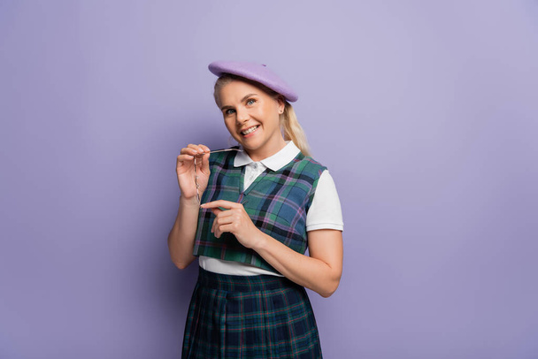 Cheerful student in uniform and beret holding eyeglasses on purple background  - Foto, afbeelding