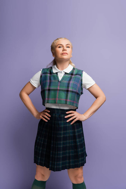 Confident student in checkered uniform holding hands on hips isolated on purple  - Foto, Bild
