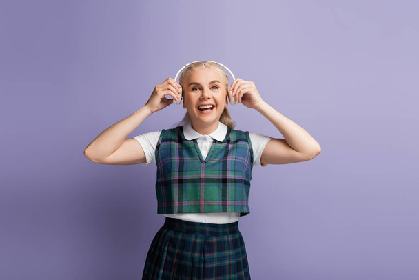 Positive student in uniform holding headphones near ears isolated on purple  - Photo, image