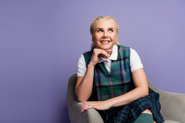 Cheerful student in checkered uniform sitting on armchair isolated on purple  - Fotografie, Obrázek