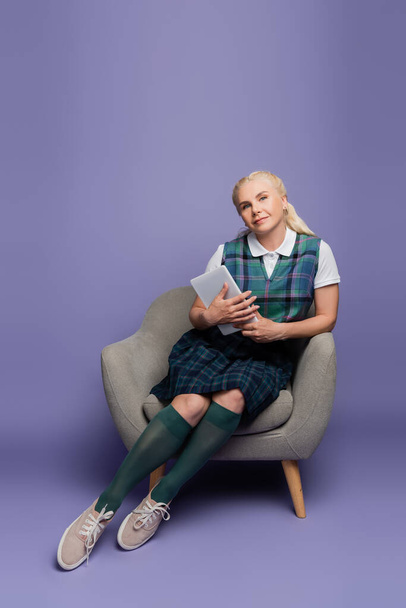 Student in plaid uniform holding digital tablet while sitting on armchair on purple background  - Foto, Imagen