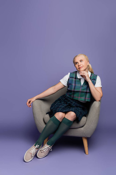 Dreamy student in plaid uniform sitting on armchair on purple background  - Fotó, kép