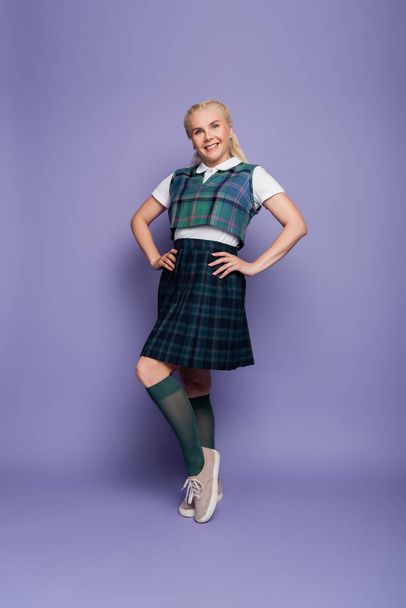 Full length of smiling student in plaid skirt standing on purple background  - Foto, immagini