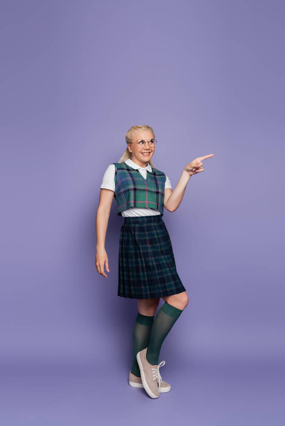 Full length of smiling student in uniform and eyeglasses pointing with finger on purple background  - 写真・画像