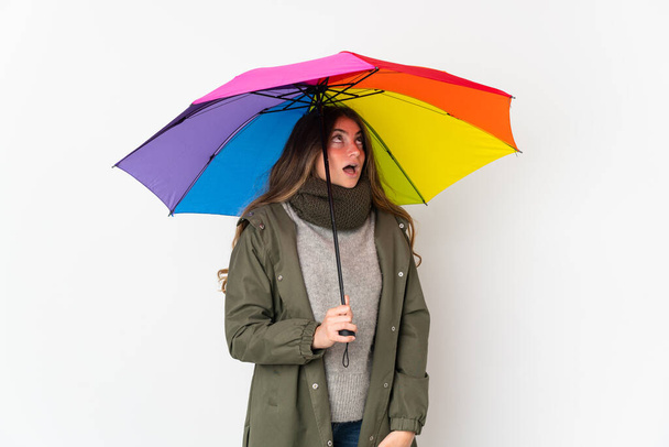 Young caucasian woman holding an umbrella isolated on white background looking up and with surprised expression - Fotografie, Obrázek
