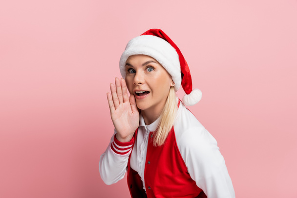 Excited woman in santa hat holding hand near cheek isolated on pink  - Photo, image