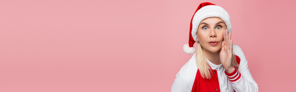 Amazed woman in santa hat holding hand near cheek isolated on pink, banner  - Fotó, kép