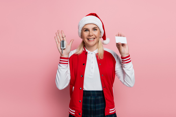 Cheerful blonde woman in santa hat holding driving license and car key on pink background - Foto, afbeelding