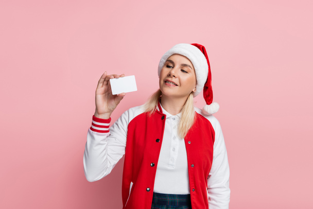 Blonde woman in red santa hat holding empty driving license isolated on pink  - Foto, afbeelding