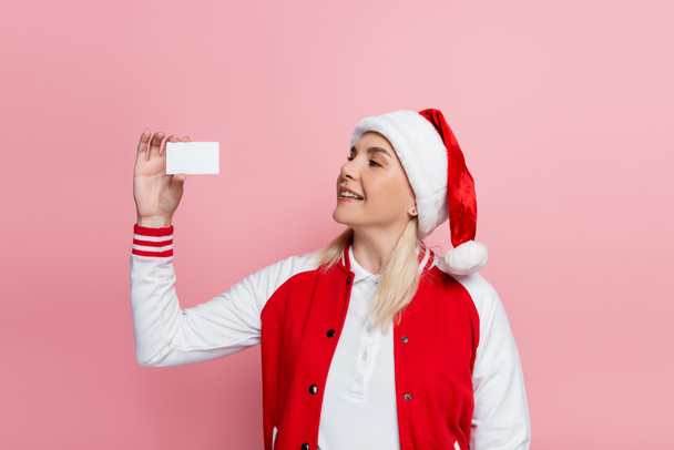 Smiling blonde woman in santa hat holding empty driving license isolated on pink  - Zdjęcie, obraz