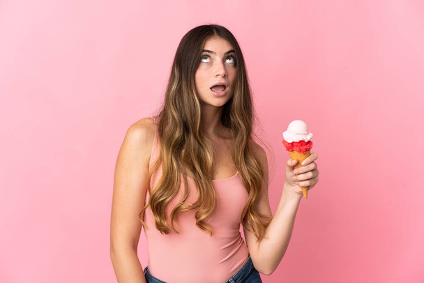 Молода біла жінка з корнетним морозивом ізольована на рожевому фоні, дивлячись вгору і з несподіваним виразом
 - Фото, зображення