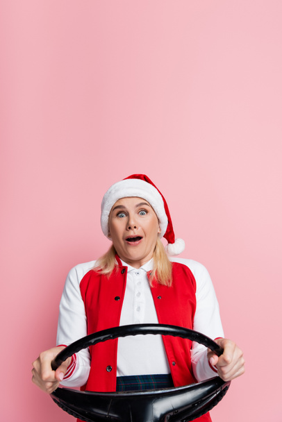 Scared woman in santa hat holding steering wheel isolated on pink  - Foto, immagini